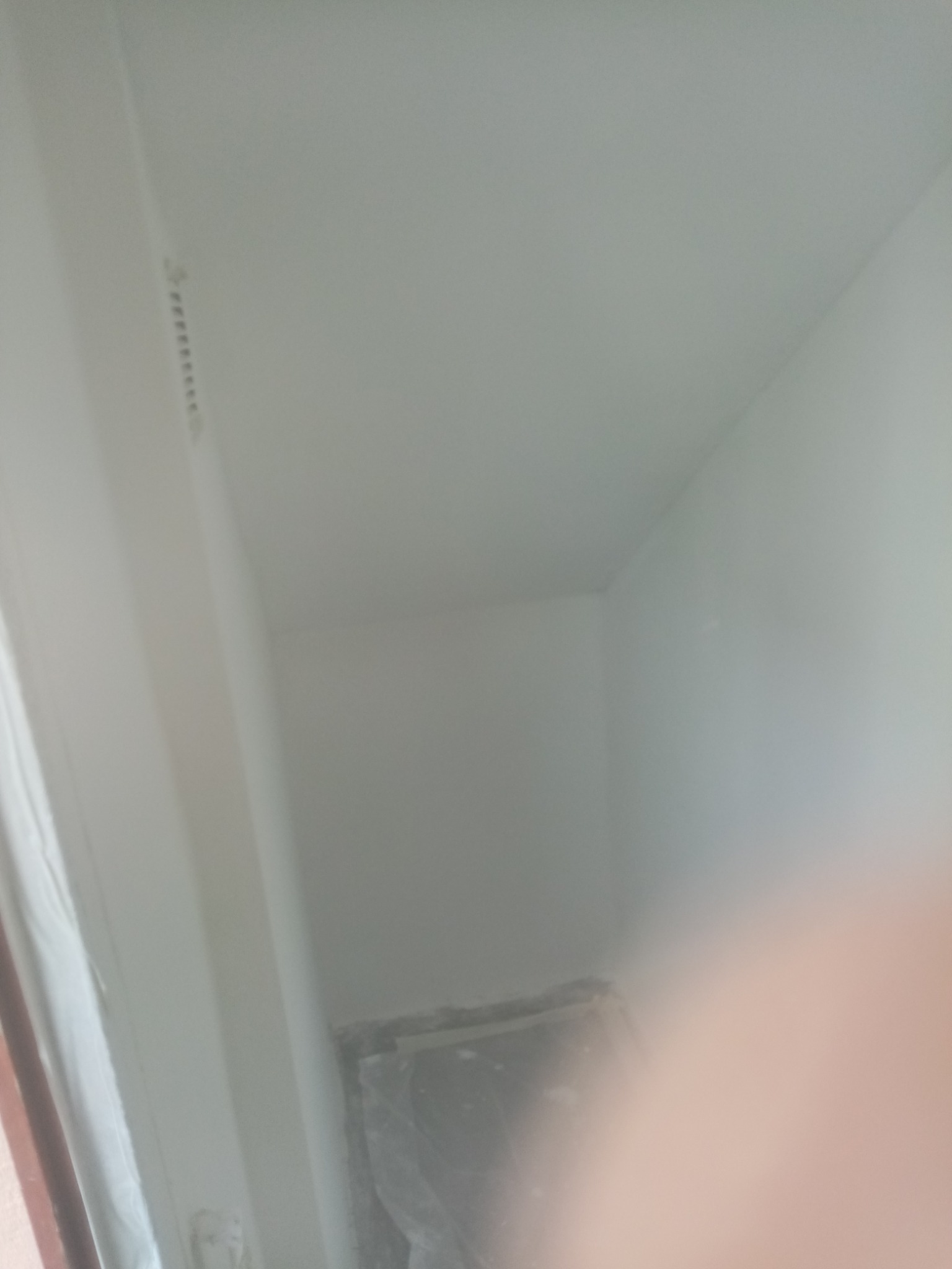 Świeżo pomalowany róg pomieszczenia z widoczną listwą karniszową i folią ochronną na podłodze, ujęcie z bliska z częściowo zasłoniętym obiektywem.