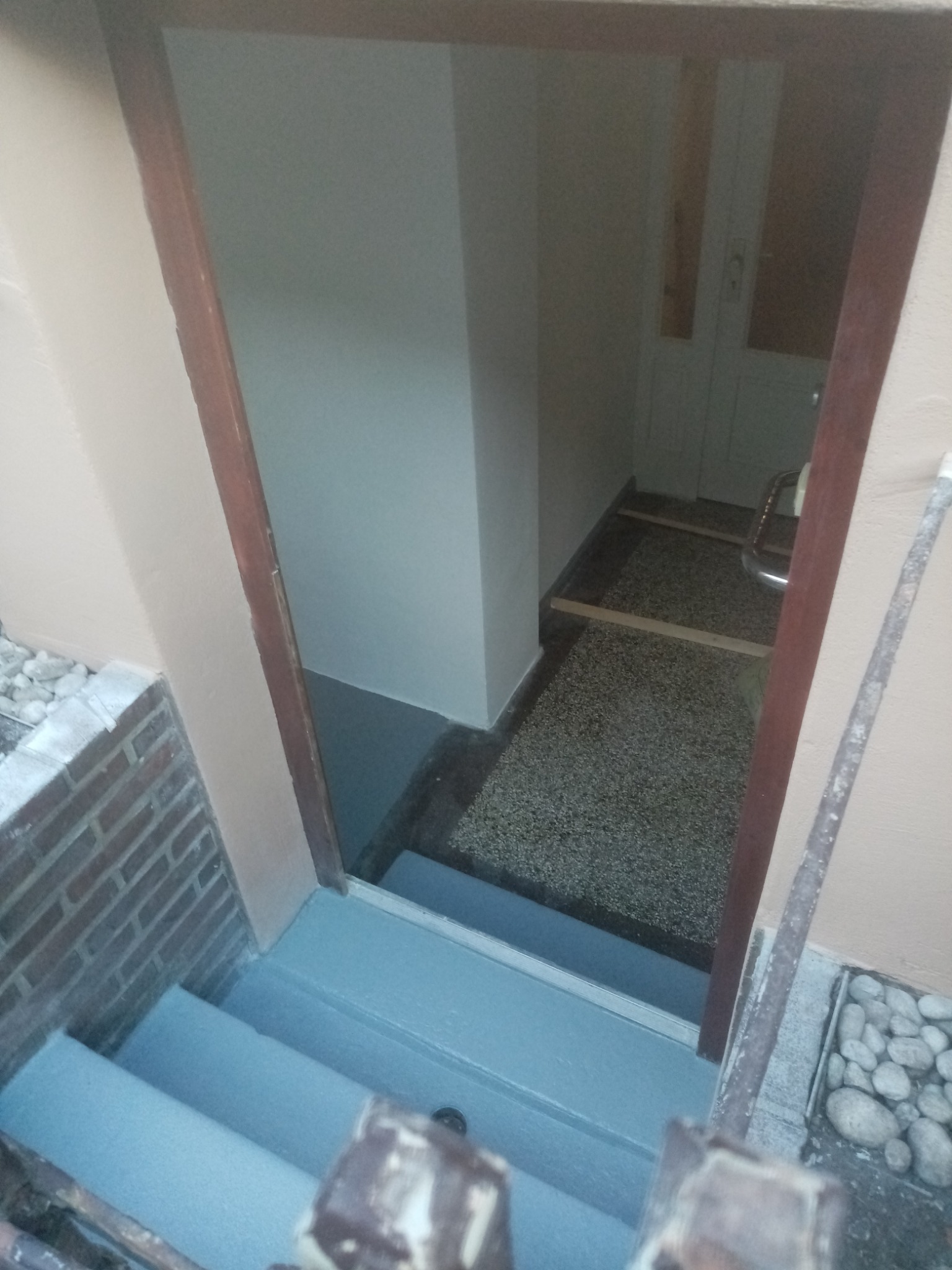 Pomieszczenie po remoncie z widocznymi pomalowanymi schodami na szaro, fragmentem ceglanej ściany i wejściem do jasnego wnętrza z widocznymi drzwiami i podłogą z lastryko.