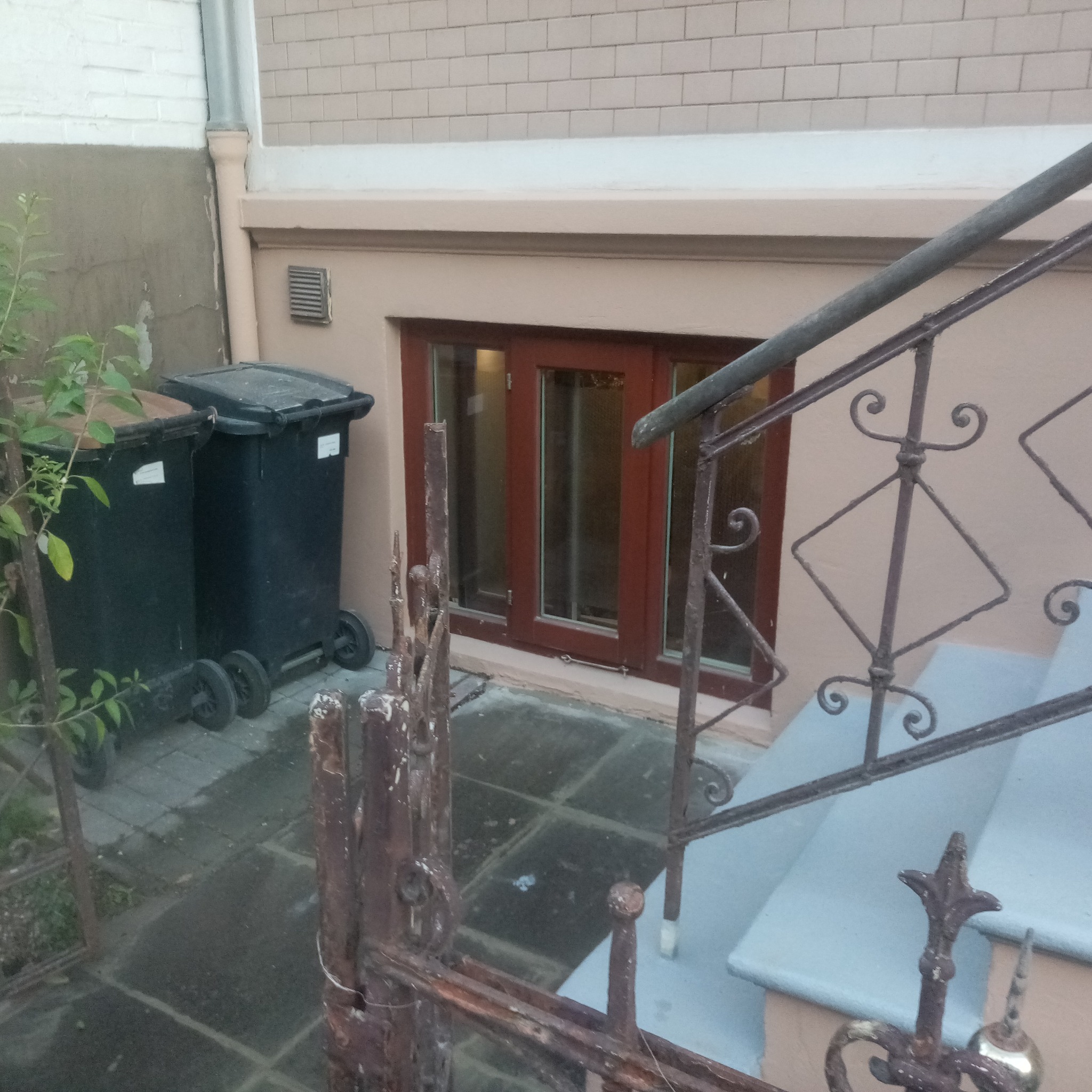 Okna piwniczne z brązowymi ramami, widoczne z zewnątrz przez ozdobną, metalową barierkę schodów. Obok stoją dwa czarne pojemniki na śmieci.