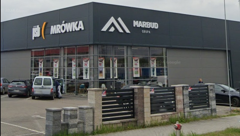 Fasada budynku z logo PSB Mrówka i Marbud Grupa, widoczny parking z samochodami i fragment ogrodzenia. Ujęcie z poziomu ulicy, w dzień.