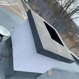 Nowo wykończony komin na dachu pokrytym papą, widok z góry na okolicę z drogą i drzewami.