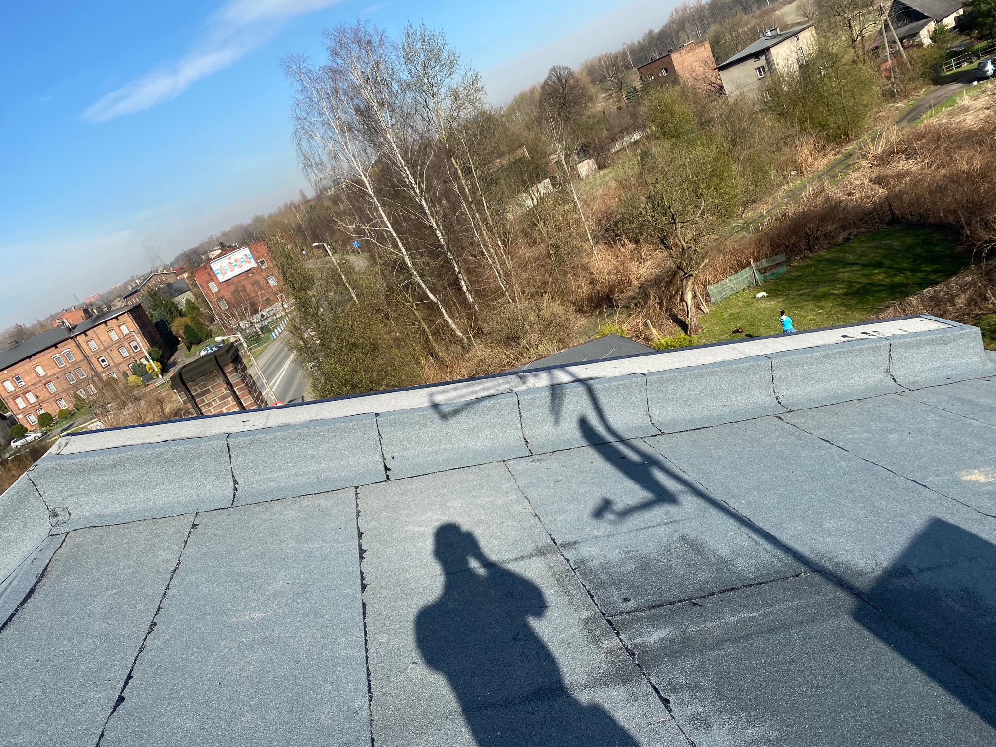 Płaski dach pokryty papą termozgrzewalną, widok z góry na okoliczne budynki i zieleń, cień osoby robiącej zdjęcie w rogu kadru.
