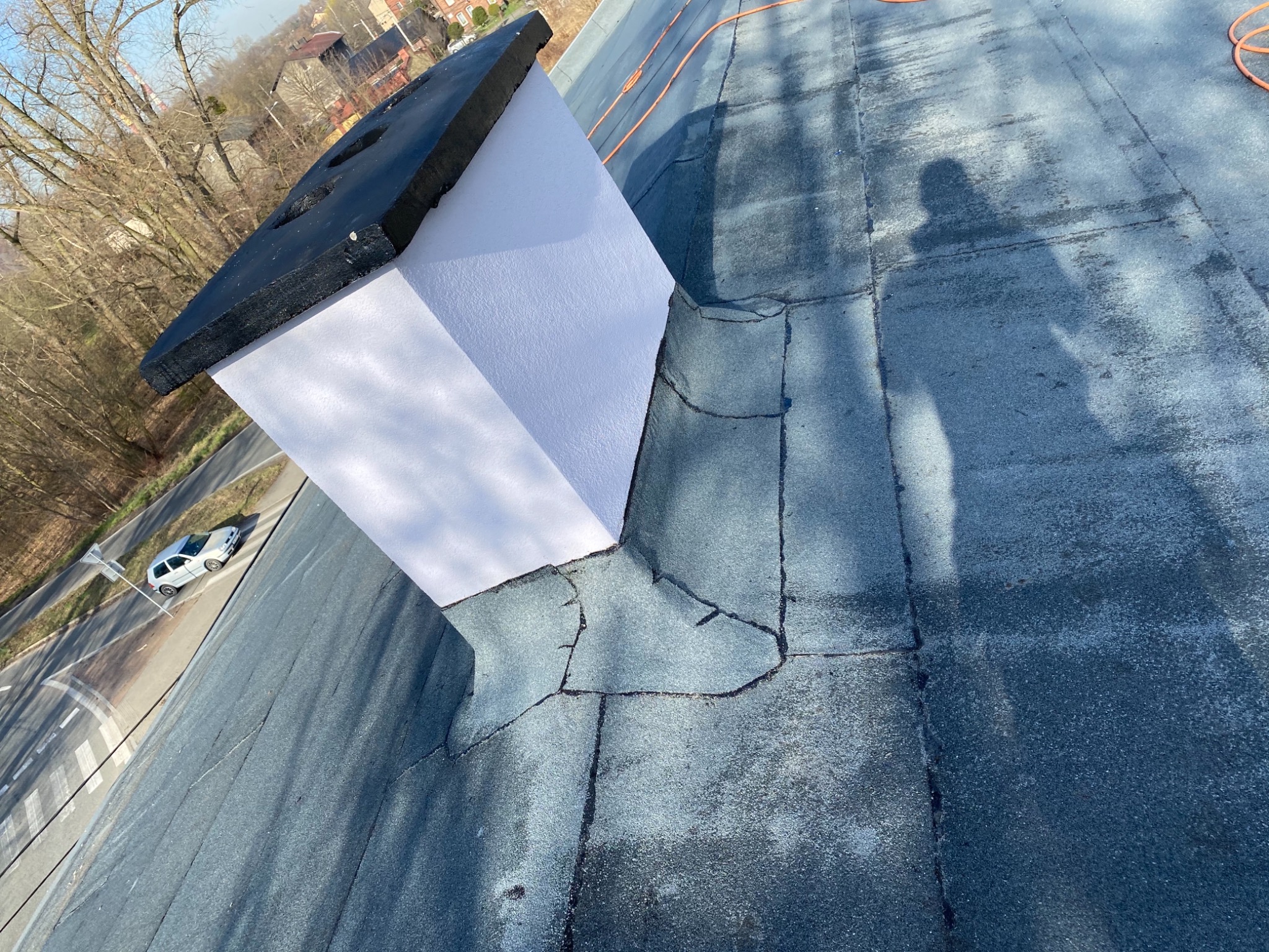 Widok z góry na płaski dach pokryty papą z widocznym kominem otynkowanym na biało i pomalowanym na czarno u góry, w tle drzewa i droga z samochodem.