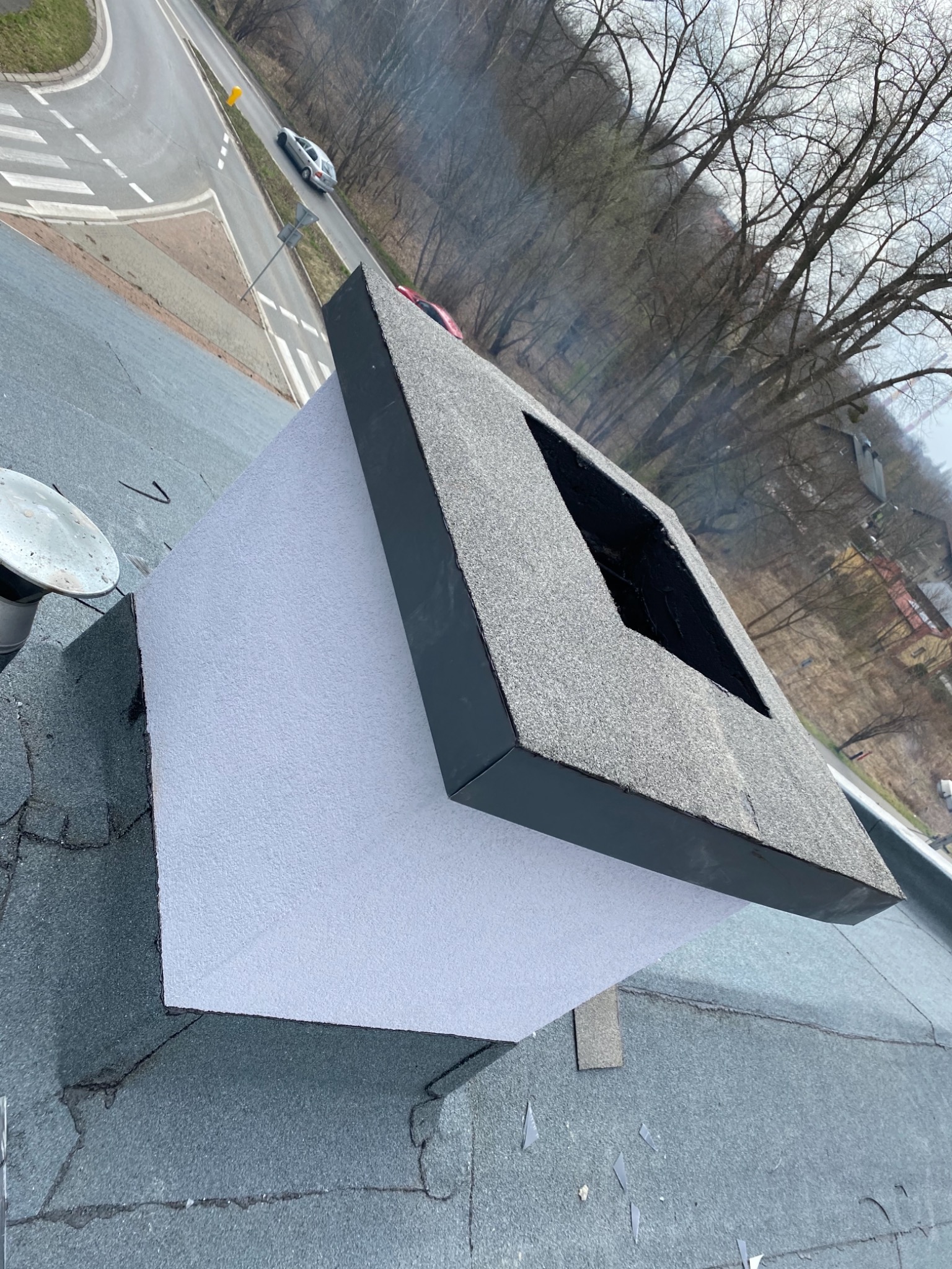 Nowo wykończony komin na dachu pokrytym papą, widok z góry na okolicę z drogą i drzewami.