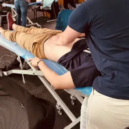 Pacjent leżący na niebieskim stole do masażu, terapeuta wykonuje masaż dolnej części pleców, inni pacjenci w tle.