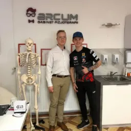 Dwaj mężczyźni pozują w gabinecie z logo 'Arcum Gabinet Fizjoterapii' na ścianie, obok szkieletu anatomicznego i umywalki.