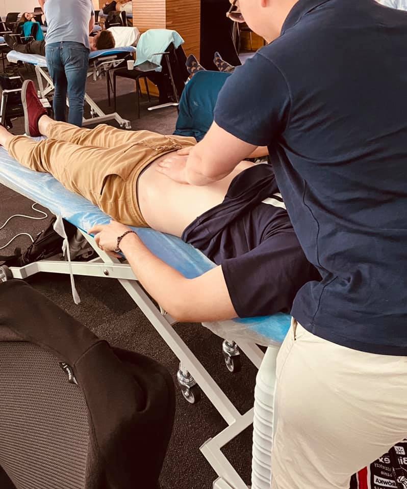 Pacjent leżący na niebieskim stole do masażu, terapeuta wykonuje masaż dolnej części pleców, inni pacjenci w tle.