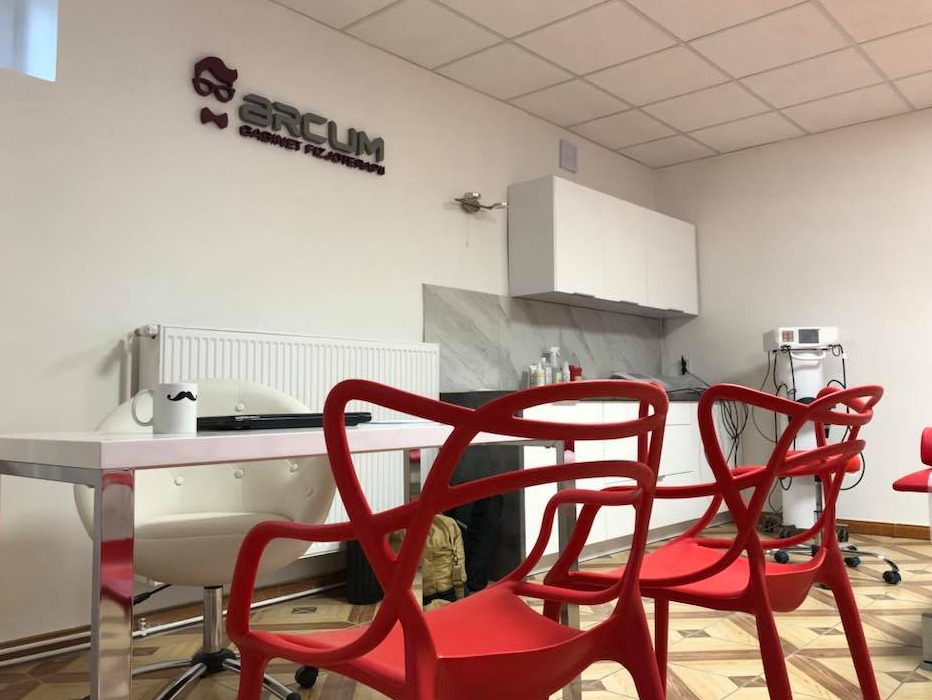 Wnętrze gabinetu z czerwonymi krzesłami, biurkiem z kubkiem z wąsami, sprzętem medycznym i logo firmy na ścianie