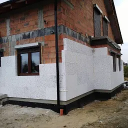 Fragment ściany domu w trakcie ocieplania styropianem, widoczne niezabezpieczone cegły na górze i fragment okna z roletą.