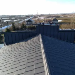 Widok z góry na nowo położony, ciemnoszary dach z blachodachówki, z widocznymi dwoma dekarzami pracującymi przy kominie na tle zimowego krajobrazu z polami i resztkami śniegu.