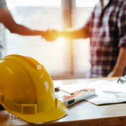 Żółty kask budowlany na stole z laptopem, poziomnicą i planami; w tle uścisk dłoni dwóch osób przy oknie.