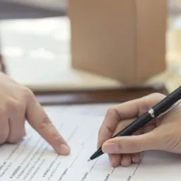 Dłoń wskazująca na fragment umowy, druga dłoń podpisuje dokument czarnym długopisem na drewnianym stole, widoczne fragmenty tekstu prawnego.