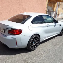 Białe BMW M2 z czarnymi felgami, widoczne z tyłu, z tablicą rejestracyjną 'MARGOT', zaparkowane na szarej nawierzchni, z pomarańczową ścianą w tle.