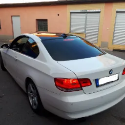 Białe BMW serii 3 Coupe z czarnym, błyszczącym dachem panoramicznym, zaparkowane na ulicy przed budynkiem z zamkniętymi roletami antywłamaniowymi.