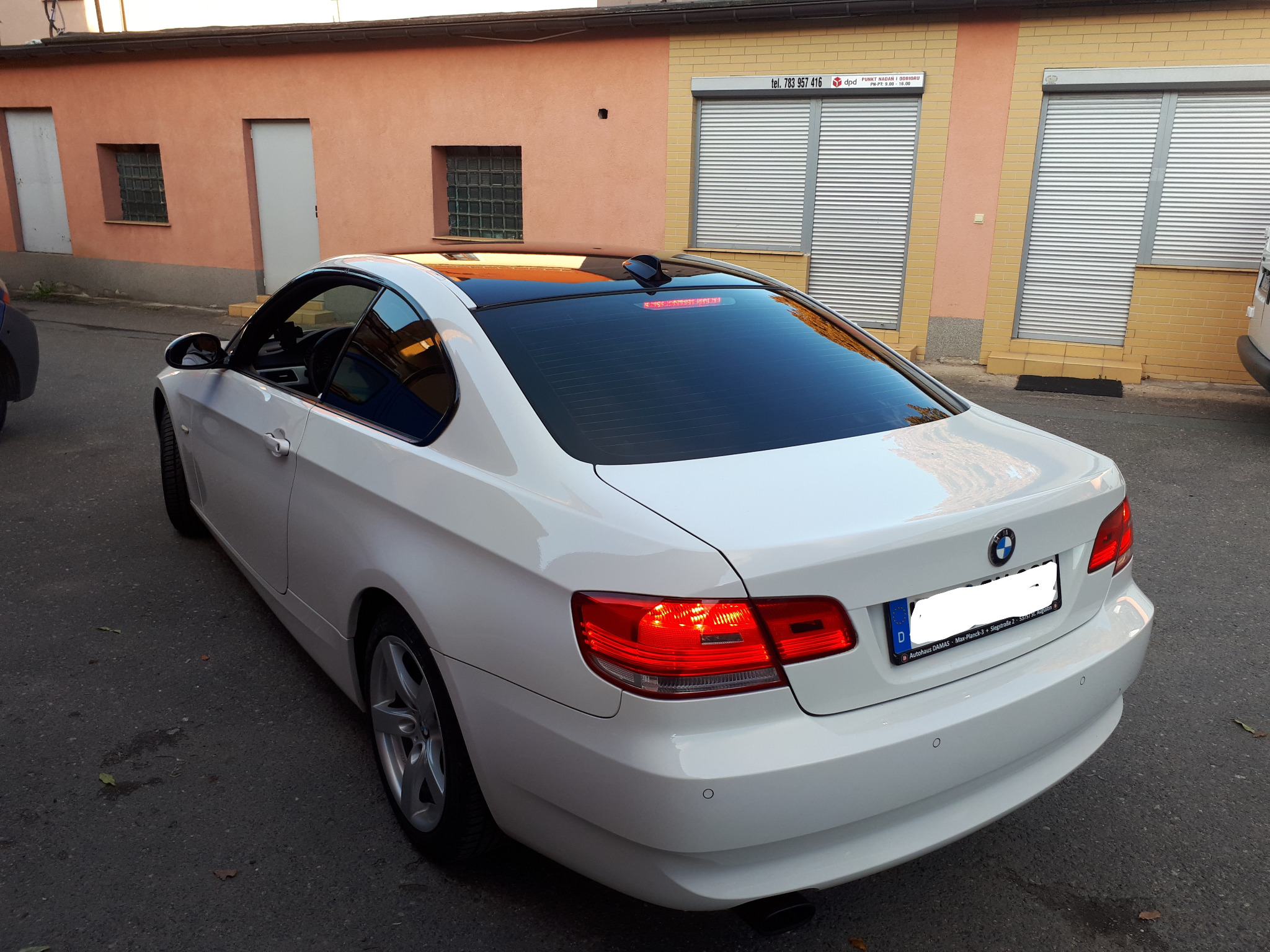 Białe BMW serii 3 Coupe z czarnym, błyszczącym dachem panoramicznym, zaparkowane na ulicy przed budynkiem z zamkniętymi roletami antywłamaniowymi.