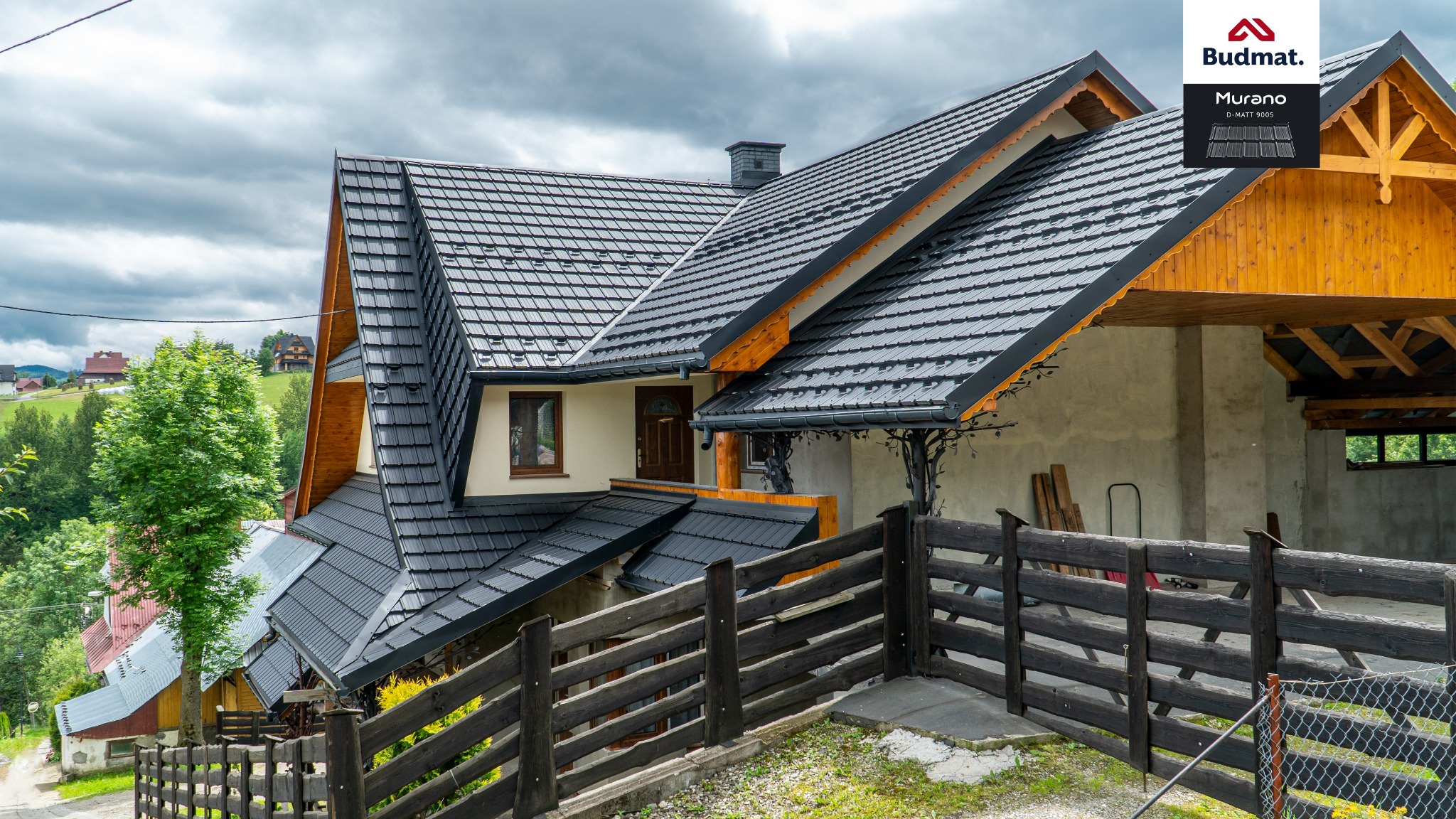 Dom z ciemną dachówką ceramiczną Murano D-Matt 9005 firmy Budmat, widok z dołu, otoczony drewnianym ogrodzeniem i zielenią, w tle zabudowa wiejska.