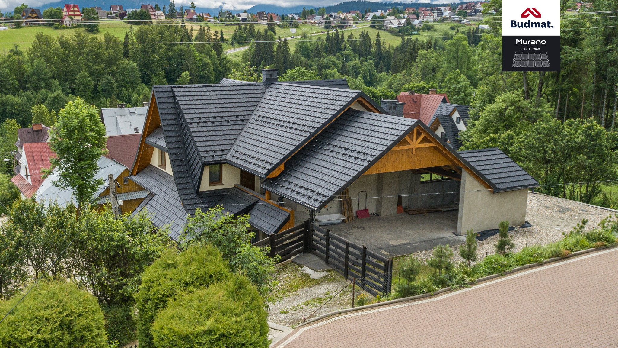 Dom jednorodzinny z ciemną, modułową konstrukcją na dachu, widok z góry na tle zieleni i zabudowań, logo Budmat w prawym górnym rogu.
