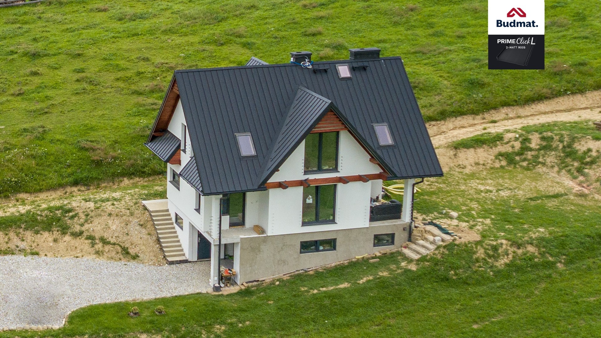 Nowoczesny dom jednorodzinny z czarnym dachem z blachy trapezowej Budmat Prime Click L, widok z góry na tle zielonego wzgórza.