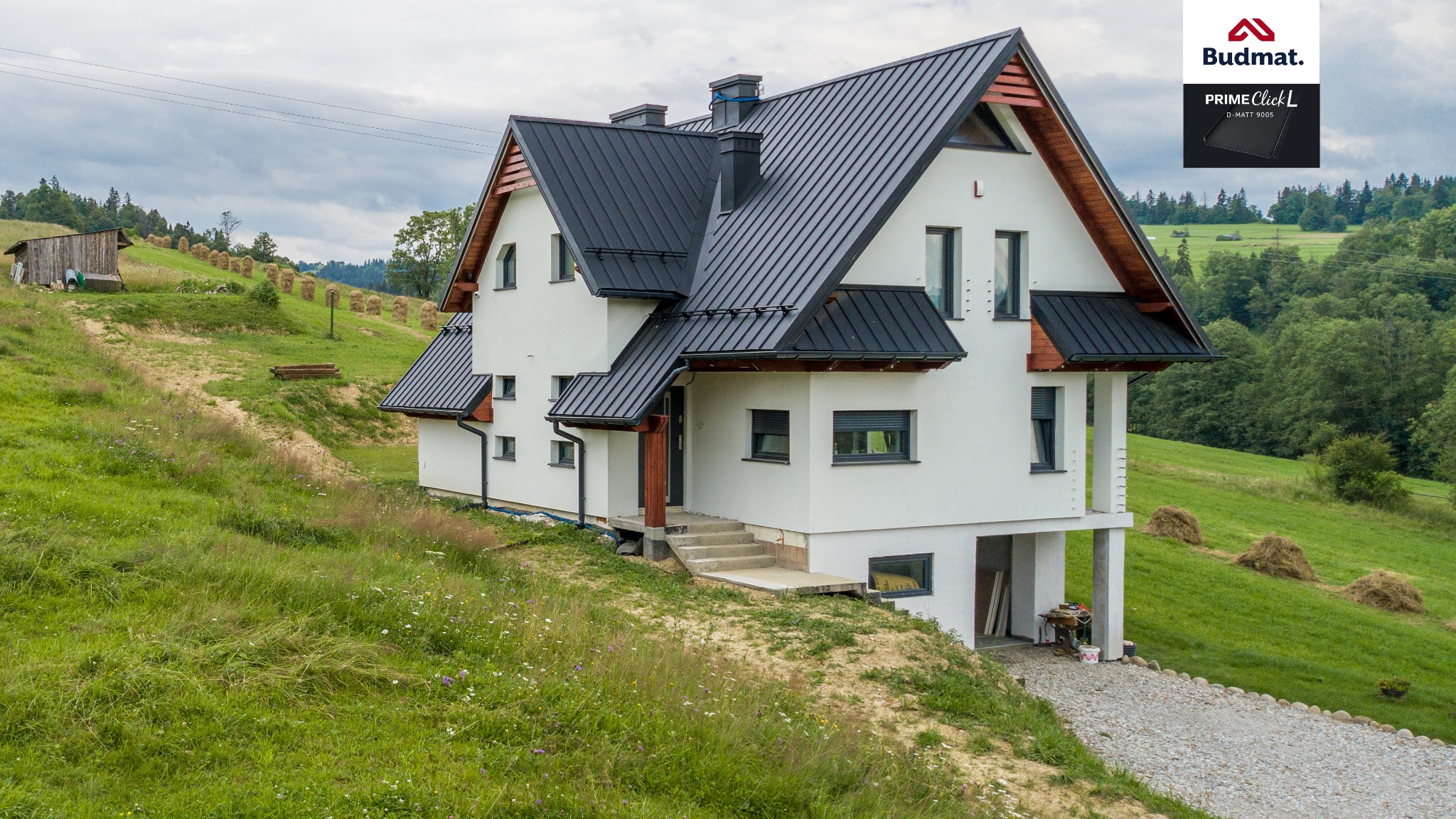 Nowoczesny dom z ciemną blachą na dachu, położony na wzgórzu z widokiem na łąki i lasy, widoczny znak firmowy Budmat.