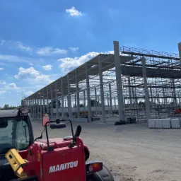 Konstrukcja szkieletowa powstającego budynku przemysłowego z betonu i stali, widoczna z perspektywy operatora czerwonego wózka widłowego Manitou na placu budowy w słoneczny dzień.