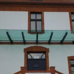 Szklany daszek nad wejściem do budynku z ceglaną elewacją, wsparty na metalowych, czarnych wspornikach.