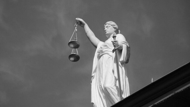 Czarno-białe zdjęcie rzeźby Temidy z wagą i mieczem na tle pochmurnego nieba, widzianej z dołu, stojącej na krawędzi dachu.