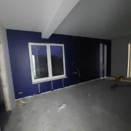 Wnętrze w trakcie prac wykończeniowych: ściany pomalowane na ciemny granat, widoczne puszki elektryczne, listwy przypodłogowe i surowa podłoga betonowa. Okno z białymi ramami w tle.