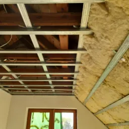 Poddasze w trakcie ocieplania wełną mineralną, widoczna konstrukcja metalowa pod płyty kartonowo-gipsowe, okno zabezpieczone folią malarską.