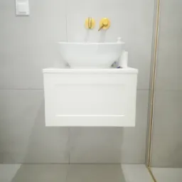 Nowoczesna łazienka z białą, podwieszaną szafką umywalkową, okrągłą umywalką nablatową i złotymi bateriami na tle szarych płytek.