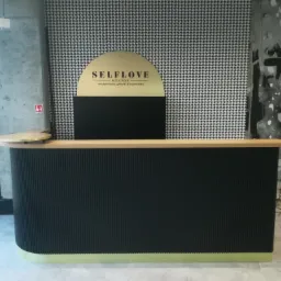 Czarna lada recepcyjna z pionowymi żłobieniami i złotymi akcentami, na tle ściany w pepitkę i logo 'SELFLOVE Gdańsk', oświetlenie górne.