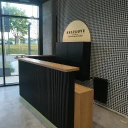Nowoczesna lada recepcyjna z czarnymi pionowymi listwami i drewnianym blatem, podświetlona u dołu zielonym paskiem, na tle ściany w pepitkę i logo firmy 'SELFLOVE Gdańsk'.