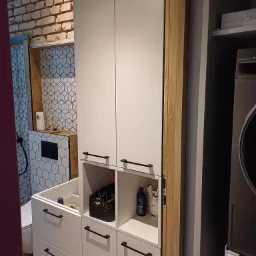 Biała szafa łazienkowa z czarnymi uchwytami na tle ściany z cegły i geometrycznych płytek, obok pralka.