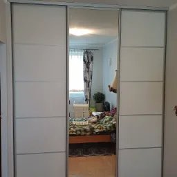 Biała szafa przesuwna z lustrem w środkowej części, odbijająca wnętrze pokoju z łóżkiem i zasłonami.