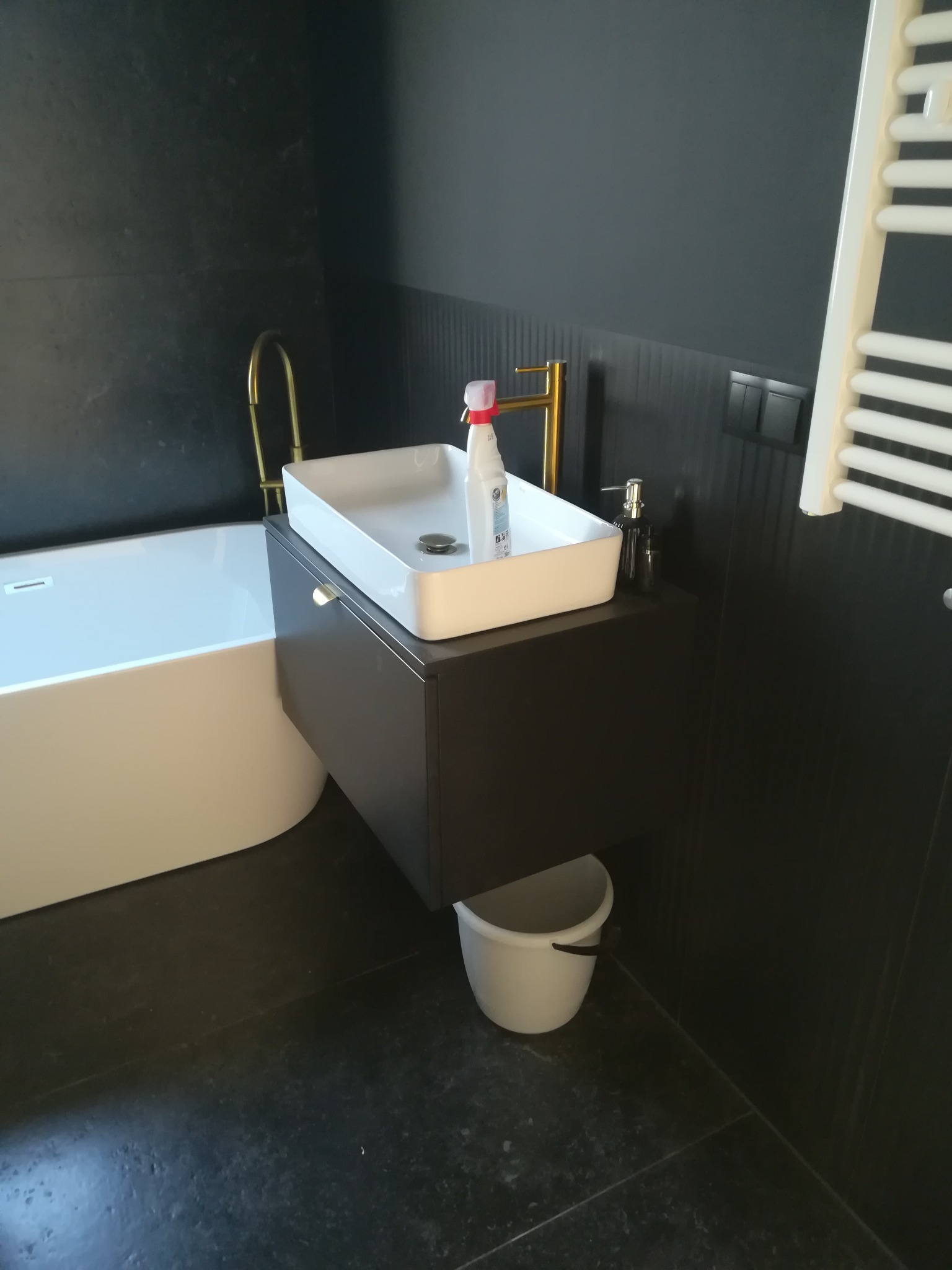 Nowoczesna łazienka z czarnymi płytkami, białą wanną i wiszącą szafką z umywalką nablatową, złota armatura i biały grzejnik drabinkowy.