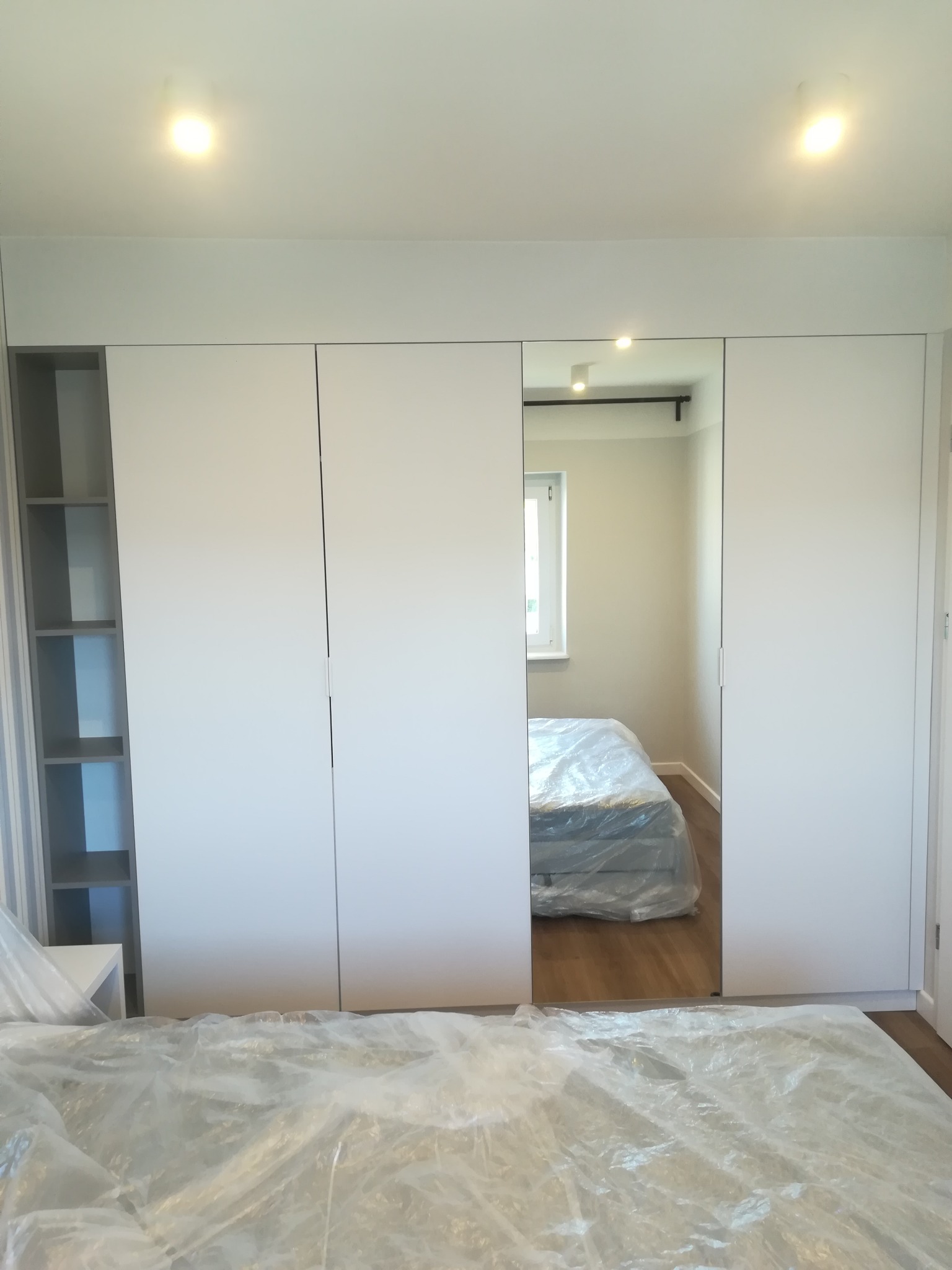 Biała szafa wnękowa z przesuwnymi drzwiami lustrzanymi, widok na łóżko zabezpieczone folią ochronną, otwarte półki z szarymi przegródkami po lewej stronie.