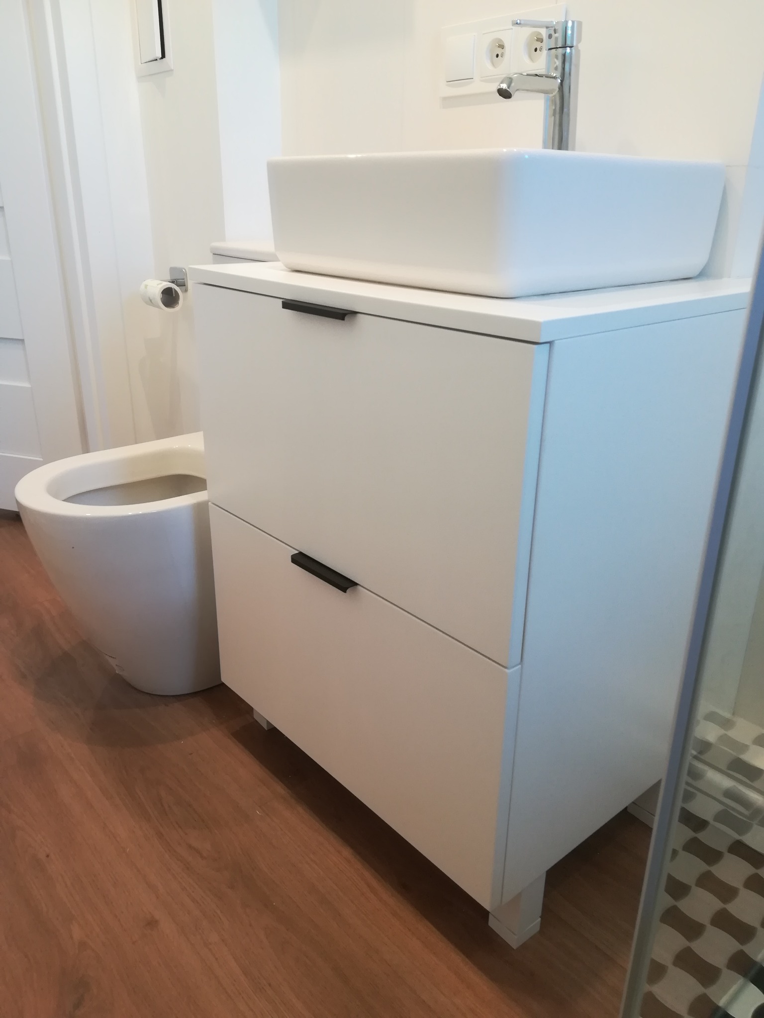 Biała szafka łazienkowa z dwiema szufladami i kwadratową umywalką nablatową, minimalistyczny design, widoczny fragment toalety i drewnopodobna podłoga.