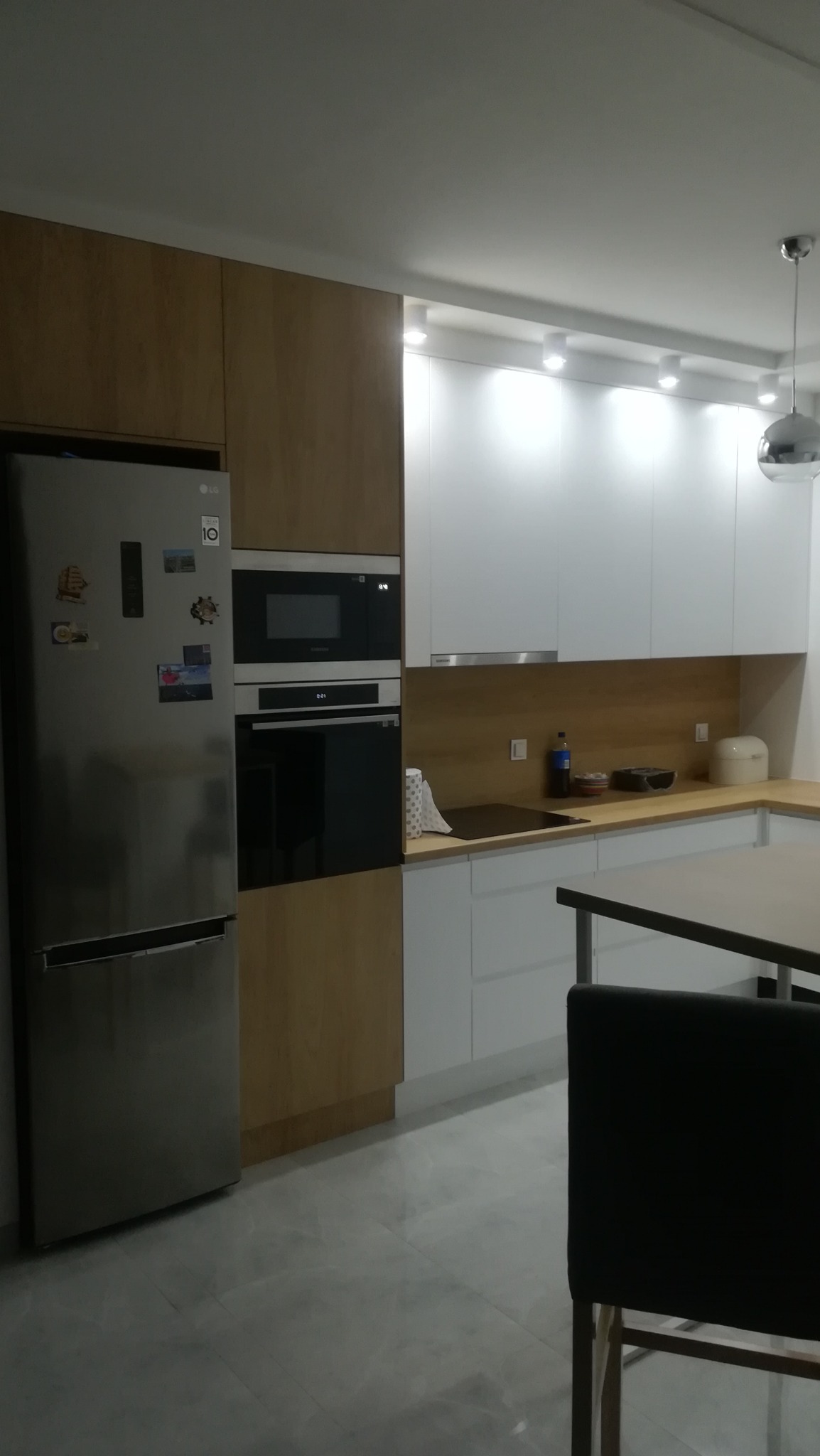 Nowoczesna kuchnia z jasnymi, drewnopodobnymi frontami szafek i białymi górnymi szafkami, wyposażona w srebrną lodówkę LG i zabudowany piekarnik z mikrofalówką Samsung.