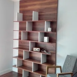 Regał na książki z forniru orzechowego z białymi przegródkami, ustawiony na tle jasnoszarej ściany, z elementami dekoracyjnymi i krzesłem w tle.