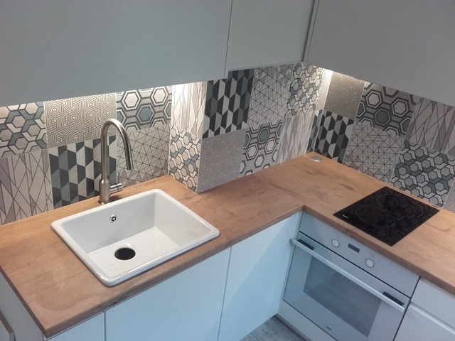 Nowoczesna kuchnia z białymi szafkami, drewnianym blatem, białym ceramicznym zlewem i płytkami ściennymi w geometryczne wzory, oświetlona listwą LED pod szafkami.