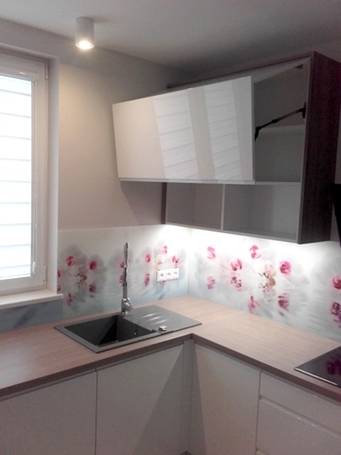 Nowoczesna kuchnia z białymi frontami, drewnianym blatem i szklanym panelem ściennym z motywem kwiatowym. Górna szafka z białym frontem podnoszona do góry.