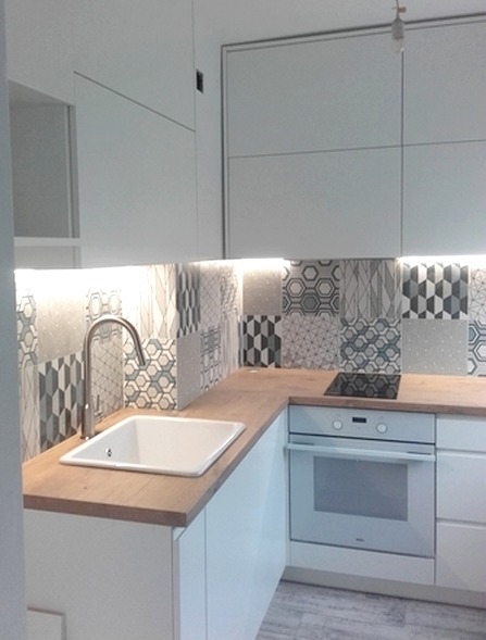 Jasna kuchnia z białymi frontami, drewnianym blatem i patchworkowym wzorem płytek nad blatem, oświetlenie LED podszafkowe.
