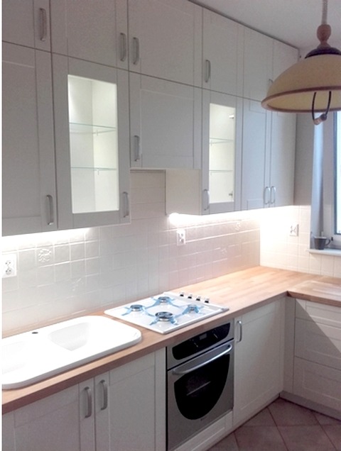 Jasna kuchnia z białymi szafkami, drewnianym blatem, ceramicznym zlewem i czteropalnikową płytą gazową, oświetlona lampą wiszącą.