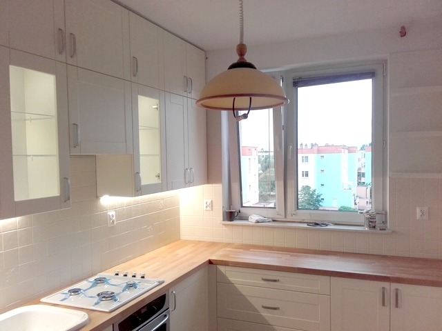 Jasna kuchnia z drewnianym blatem, białymi szafkami górnymi z przeszkleniami i oświetleniem podszafkowym. Widoczna płyta gazowa i okno z widokiem na budynki mieszkalne.