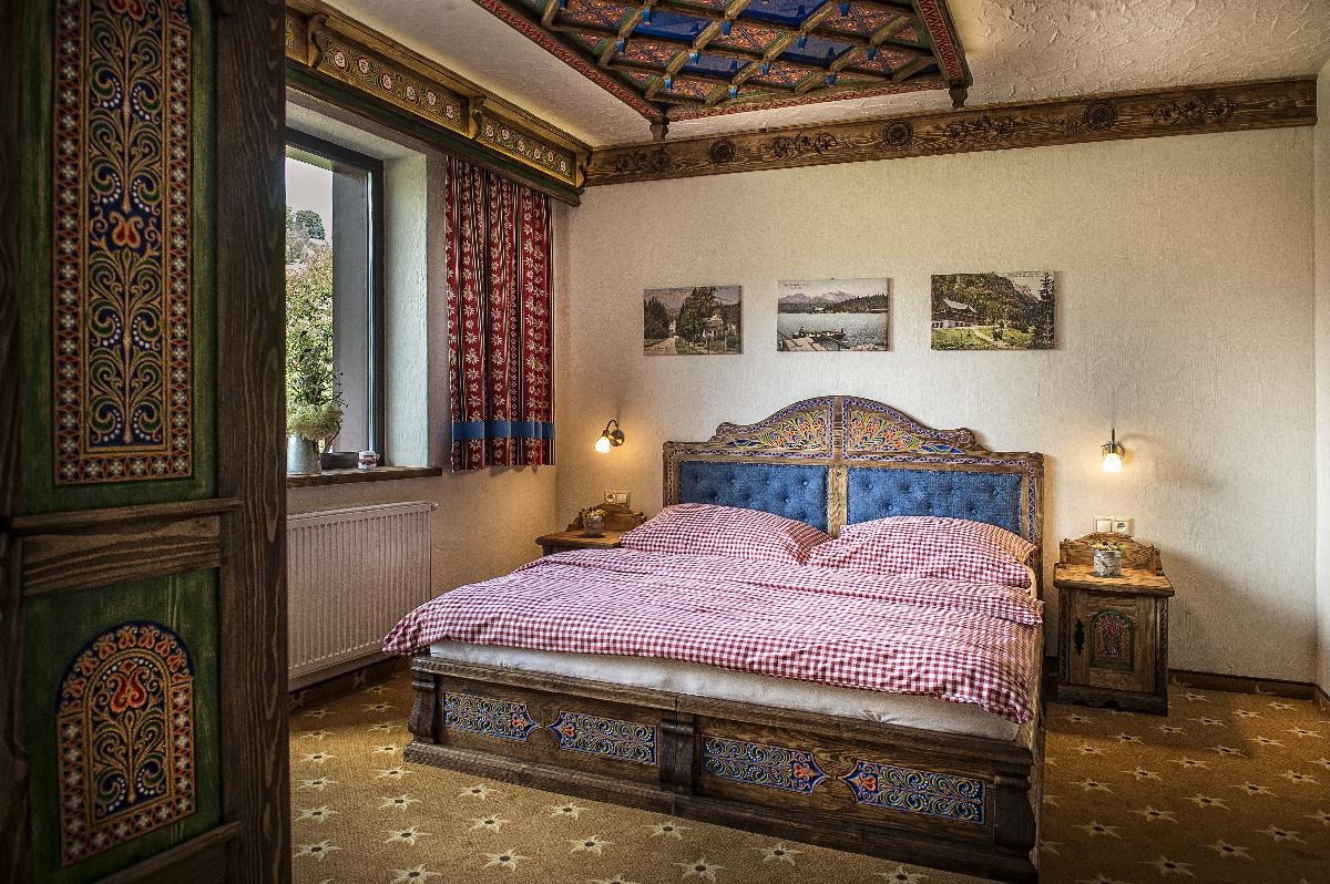 Pokój z łóżkiem w stylu rustykalnym, bogato zdobionym drewnianym wezgłowiem i ramą, czerwono-białą pościelą w kratę, wzorzystym dywanem i oknem z widokiem na zieleń.