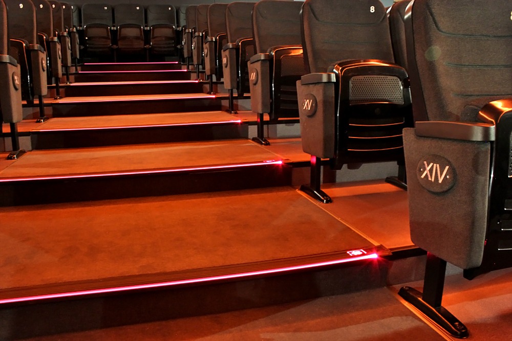 Rzędy foteli kinowych w ciemnej tonacji z numeracją 'XIV' i '8' na podłokietnikach, podświetlone schody z subtelnym, czerwonym oświetleniem LED.