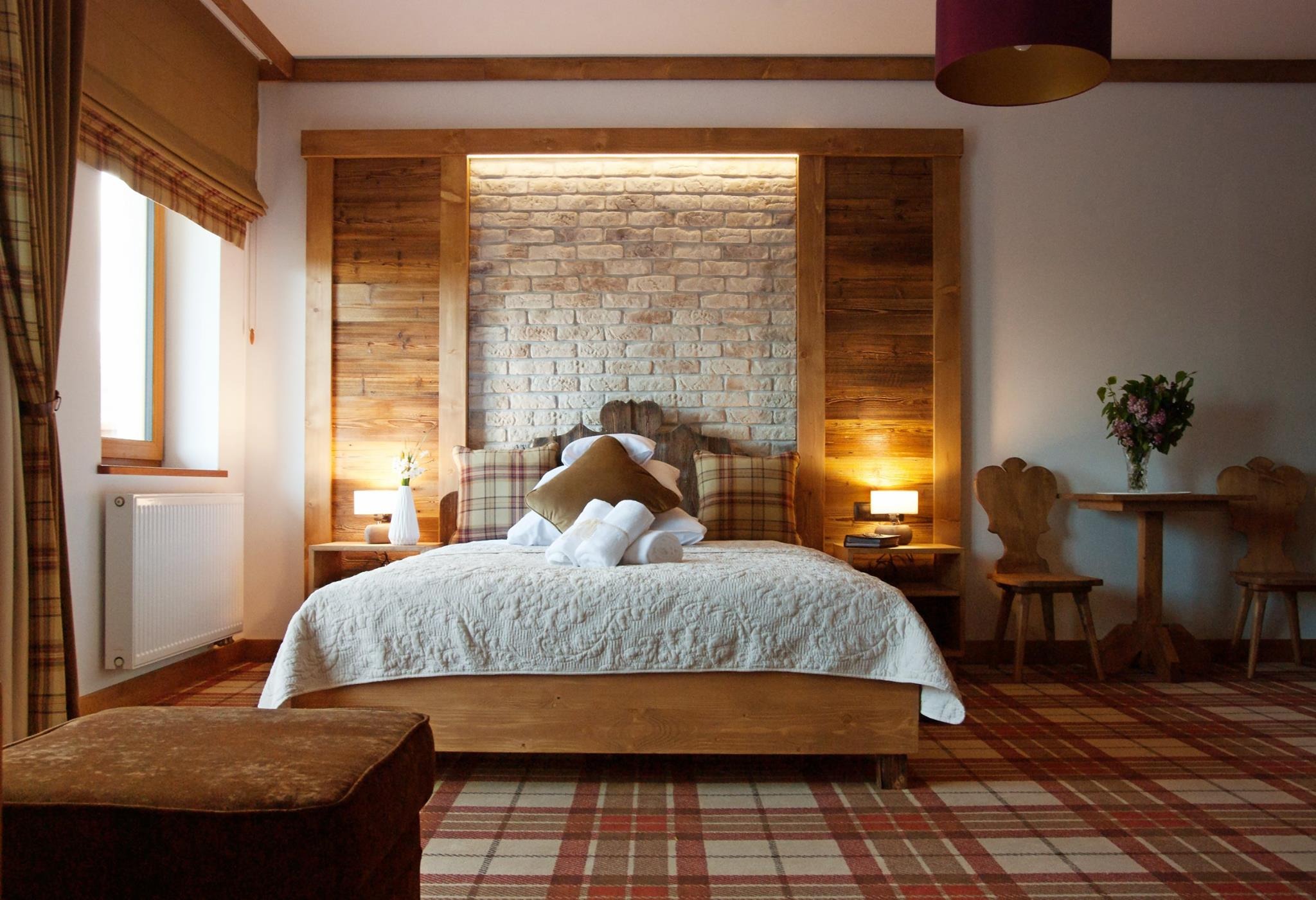 Rustykalna sypialnia z drewnianą ramą łóżka, ceglanym wezgłowiem podświetlonym LED, wzorzystą wykładziną podłogową i drewnianymi krzesłami przy stoliku z bukietem kwiatów.