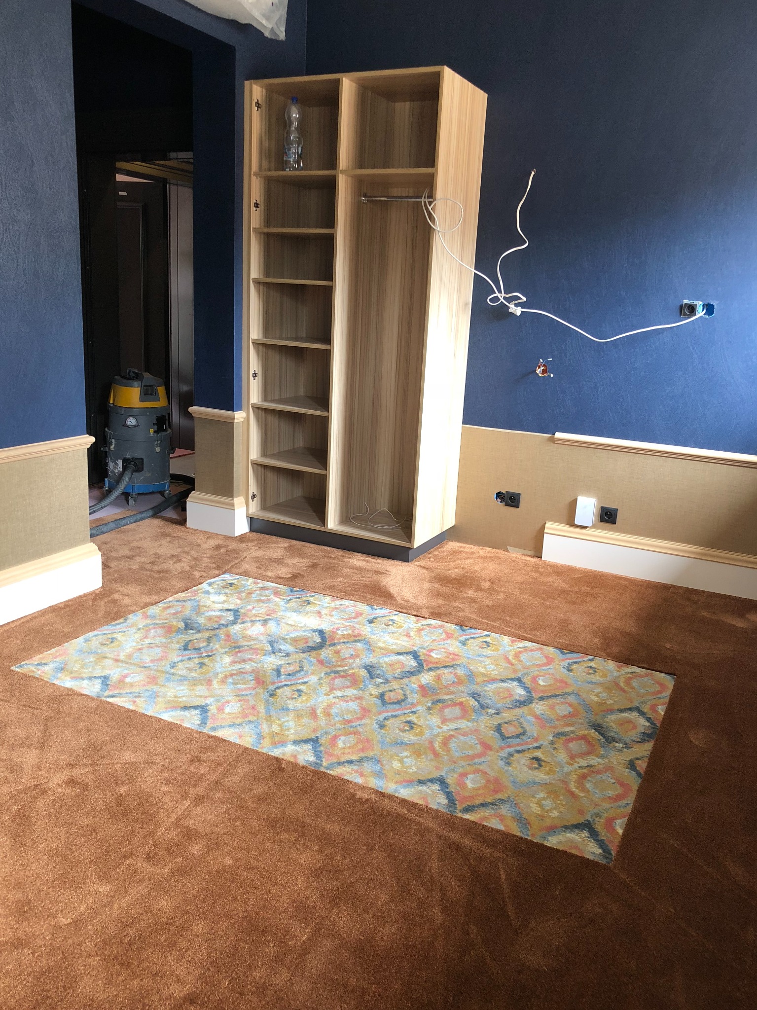 Pokój w trakcie wykańczania z brązową wykładziną dywanową i wzorzystym dywanem, widoczny odkurzacz przemysłowy, szafa z półkami i niedokończone gniazdka elektryczne na niebieskiej ścianie.