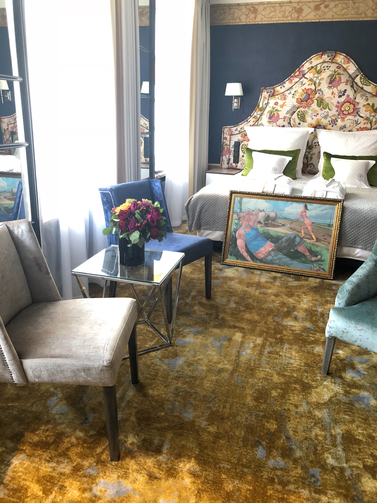 Luksusowy pokój hotelowy z wzorzystą wykładziną w odcieniach złota, z łóżkiem z kwiatowym zagłówkiem, stolikiem z bukietem róż i obrazem opartym o łóżko.