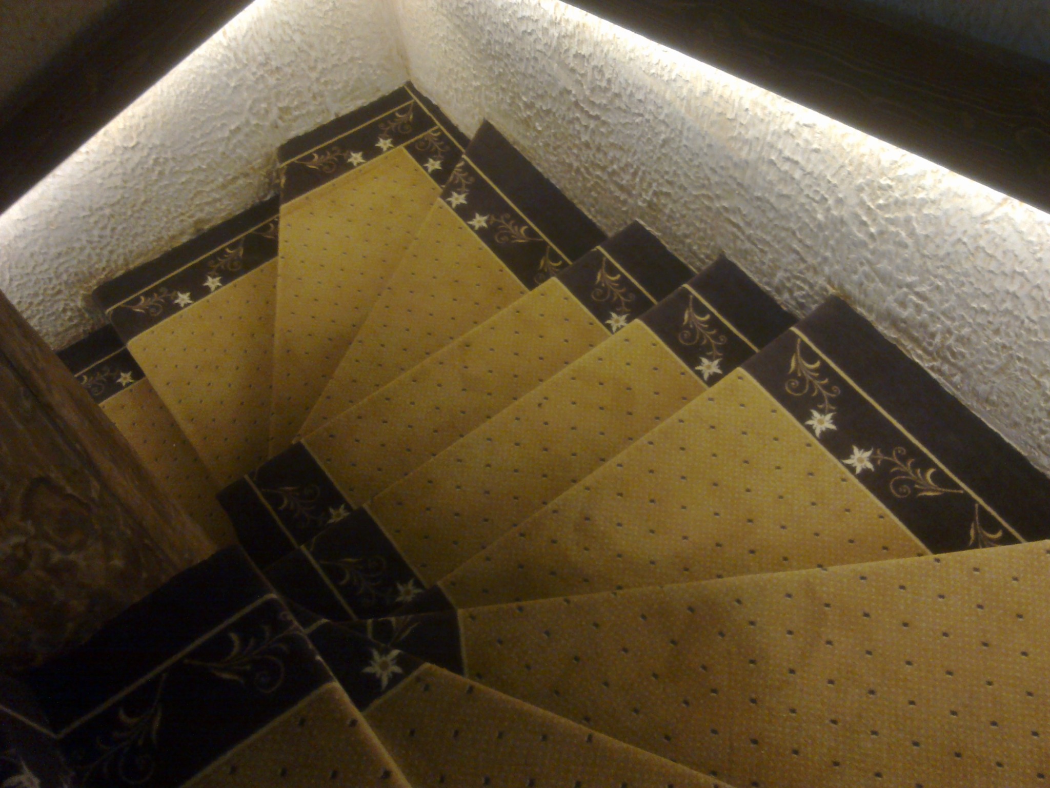 Widok z góry na schody pokryte żółtą wykładziną w drobne kropki, z ciemnobrązowymi brzegami ozdobionymi motywem gwiazd i roślinnych ornamentów; ściana obok schodów ma fakturę przypominającą baranek...