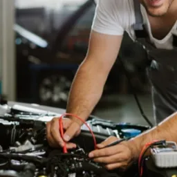 Diagnostyka silnika samochodowego: mężczyzna w kombinezonie roboczym używa multimetru do sprawdzenia instalacji elektrycznej w komorze silnika z otwartą maską.